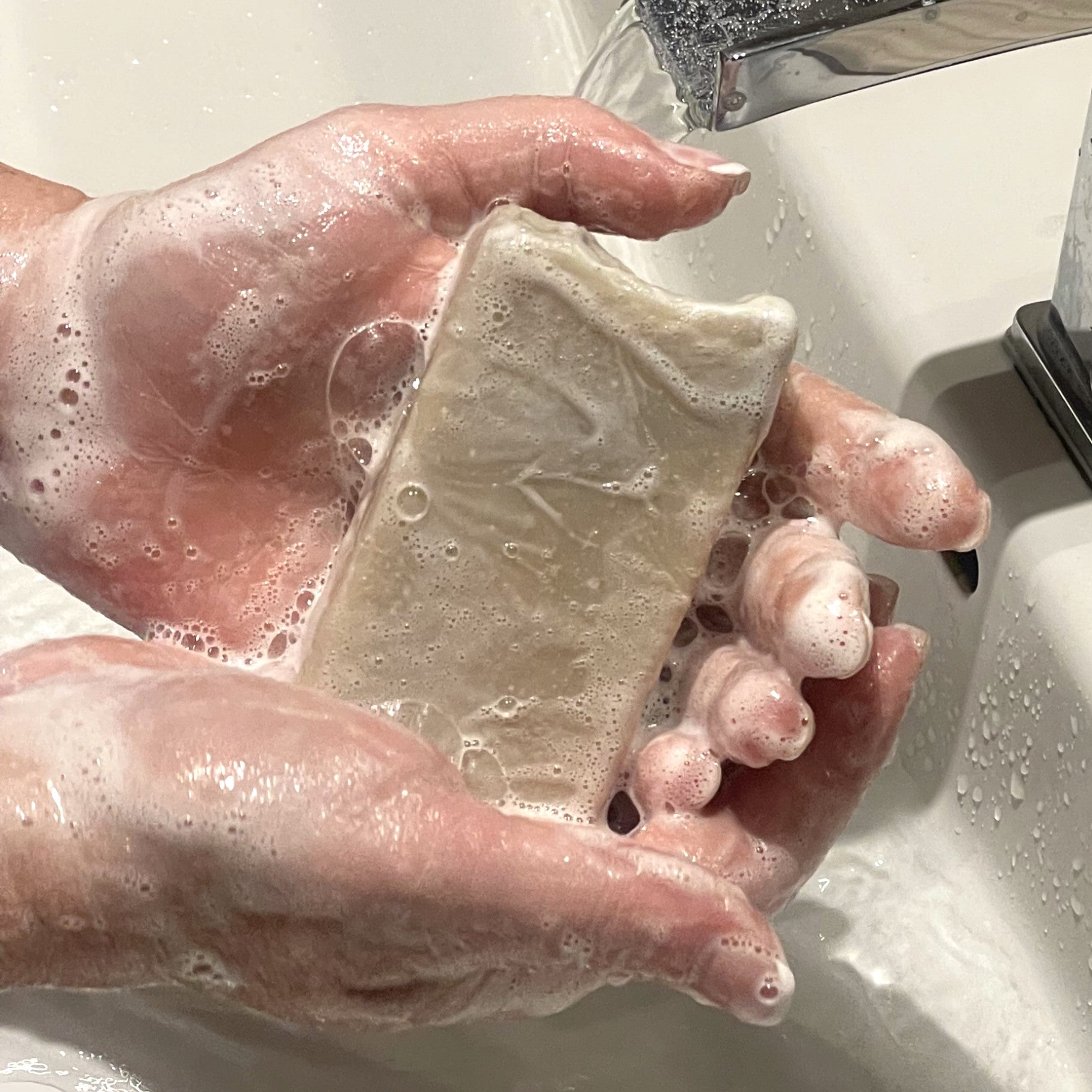 mains qui tiennent un savon rectangulaire crème couvert de mousse au dessus d'un lavabo